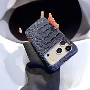 May include: A dark blue phone case with a crocodile skin texture. The case has a glossy finish and features a camera module with three lenses. The case is designed to fit a smartphone.