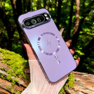 Può includere: Una custodia per telefono color lavanda con un proteggi obiettivo argentato e un cerchio di ricarica MagSafe argentato. La custodia ha un bordo argentato metallico e un design argentato decorativo nella parte inferiore. Lo sfondo è una vista sfocata di fogliame verde.