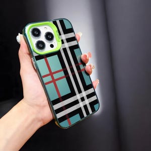 May include: A teal and red plaid phone case with a green camera lens protector. The case has a black and white plaid pattern overlaying the teal background. The camera lens protector has the text "SO COOL" printed on it.