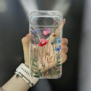 Könnte beinhalten: Transparente Handyhülle mit floralem Design. Die Hülle zeigt rote, rosa und blaue Blumen auf transparentem Hintergrund. Die Blumen sind in einem feldartigen Muster angeordnet, mit grünen Stielen und Blättern.