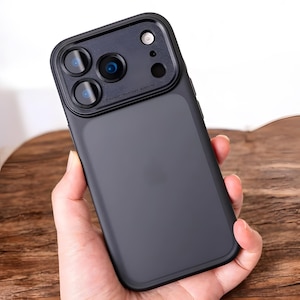 Pode incluir: Uma capa de telefone preta fosca com protetor de lente. A capa tem um acabamento liso e apresenta uma seção elevada ao redor das lentes da câmera. O protetor da câmera tem três lentes e um flash.