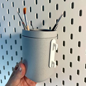 Może przedstawiać: Jasnoszary, nakrapiany, cylindryczny pojemnik do przechowywania z białym klipsem montażowym. Pojemnik mieści kilka pędzli z pomarańczowym i czarnym włosiem. Pojemnik jest przeznaczony do zawieszenia na tablicy perforowanej.