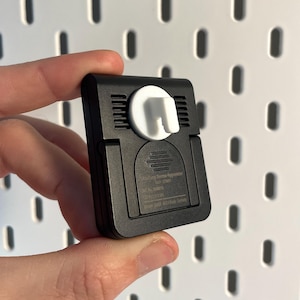 Puede incluir: Un dispositivo rectangular negro con un accesorio blanco y curvo. El dispositivo tiene ranuras horizontales y parece ser un sensor de temperatura o humedad. Se sostiene contra un fondo blanco de tablero perforado.