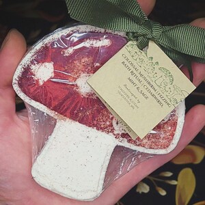 May include: A mushroom-shaped bath bomb, red and white, wrapped in clear plastic. The bath bomb has a tag with the text "COLOSSAL MUSHROOM | FIZZING BATH RITUAL | CEDARWOOD, MINT & SAGE". A green ribbon is tied around the package.