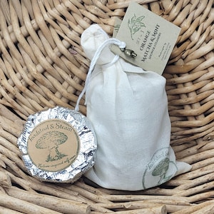 May include: A wicker basket contains a white drawstring bag and a foil-wrapped item. The bag has a tag that reads "Orange Matcha & Mint Shower Steamers." The foil-wrapped item has a label that says "Toadstool & Steam, Nature-inspired self-care."