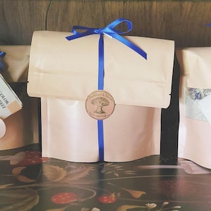Puede incluir: Varias bolsas de papel beige atadas con cintas azules, cada una con una etiqueta y una pequeña cuchara de madera. Las bolsas parecen contener productos de baño. Una pegatina redonda con un diseño de árbol está en la bolsa central.