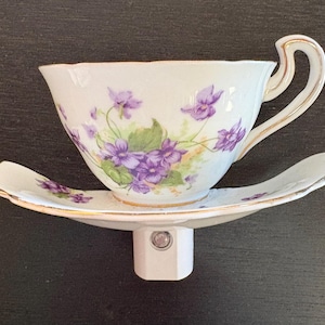 May include: A white porcelain teacup and saucer night light with a floral violet design. The cup and saucer have gold trim. The night light base is white and has a simple, modern design.