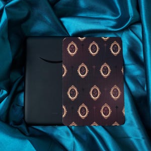 Puede incluir: Un lector de libros electrónicos negro con el logotipo de Amazon, junto a una funda decorativa. La funda presenta un patrón repetido de retratos ovalados con marco dorado sobre un fondo oscuro. Los artículos se exhiben sobre tela de satén turquesa.