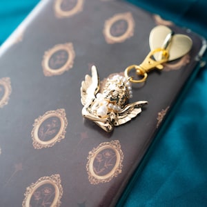 May include: A gold-colored angel charm with pearl accents and a heart-shaped clasp. The charm is attached to a dark brown notebook with gold-framed floral designs. The background is a teal fabric.