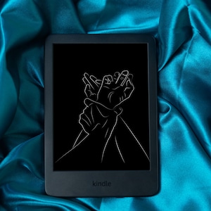 Puede incluir: Un lector de libros electrónicos Kindle negro con una pantalla negra que muestra un dibujo lineal blanco de dos manos entrelazadas. El lector de libros electrónicos está sobre una tela de satén turquesa. La palabra "kindle" está impresa en la parte inferior del dispositivo.