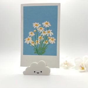 May include: A floral art print featuring white daisies with yellow centers against a light blue background. The print is held by a white cloud-shaped holder with a smiling face. The overall aesthetic is whimsical and cheerful.