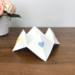 May include: A white paper fortune teller with colorful heart accents. The fortune teller is folded in a zig-zag pattern, with a yellow, blue, and pink heart on each of the four visible flaps. The object is sitting on a wooden surface.