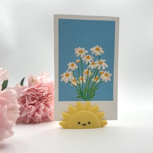 Puede incluir: Imagen de temática floral con una tarjeta azul y blanca con una ilustración de margaritas, sostenida por un soporte amarillo en forma de sol. Claveles rosas en primer plano crean una composición alegre y brillante.