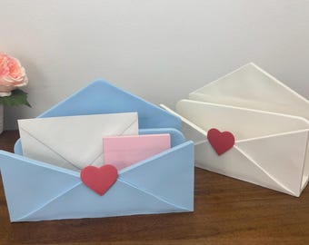 May include: Two envelope-shaped holders, one light blue and one white, each featuring a red heart-shaped accent on the front flap. The light blue holder contains a white envelope and a small pink rectangular note card inside its open compartment.