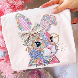 May include: A white fabric item featuring an embroidered bunny design. The bunny has floral patterned ears, a bow, and round glasses. The design incorporates pink, blue, and green colors, with pearl accents around the bunny's face and ears.
