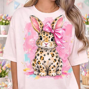 以下が含まれることがあります： ヒョウ柄の模様と大きなピンクのリボンが付いたウサギの気まぐれなイラストが特徴の、明るいピンク色のTシャツ。 ウサギはピンクとカラフルな背景に配置され、遊び心のある芸術的なデザインになっています。