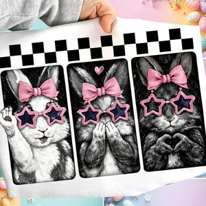May include: A white sheet featuring three black and white illustrations of rabbits. Each rabbit wears pink star-shaped sunglasses and a pink bow. The top of the sheet has a black and white checkered pattern.