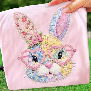May include: A pink fabric item with a patchwork bunny face wearing heart-shaped glasses. The bunny has floral patterned ears and a pink nose. The design is decorated with pearls and small pink roses.
