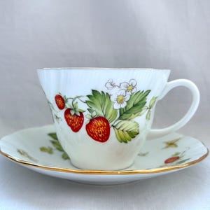 May include: White teacup and saucer set with a strawberry design. The cup and saucer feature red strawberries, green leaves, and white flowers. The rims are trimmed with gold.