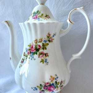 May include: A white porcelain coffee pot with a lid, handle, and spout. The pot is decorated with pink and blue floral patterns and gold trim. The design is a classic, elegant style.