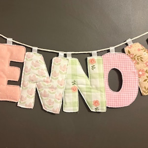 May include: A fabric banner spelling "LENNON" with individual letters in various floral and patterned fabrics. The letters are suspended from a white string with small white ribbons. The background is a solid dark gray.