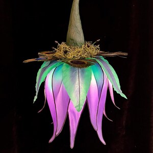 May include: A hanging decorative ornament shaped like a flower with long, pointed petals in shades of purple and teal. The top features a textured, cone-shaped stem, a cluster of dried moss, and several peacock feathers arranged in a circular pattern.
