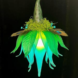 May include: A hanging light fixture featuring a pointed, textured gray top, a ring of bright green moss with small blue beads, and a layer of peacock feathers. The lower section consists of layered green and teal leaves surrounding a glowing light.