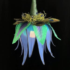 May include: A hanging light fixture shaped like a large flower with blue, translucent, downward-curving petals. The top features green leaves, peacock feathers, and a textured green stem. A small, glowing light source is visible inside the petals.