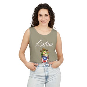 Puede incluir: Camiseta sin mangas verde oliva claro con la palabra "Latina" en escritura cursiva. La camiseta sin mangas presenta una rana de dibujos animados tocando un tambor con el diseño de la bandera de Puerto Rico. La rana lleva un sombrero de paja.