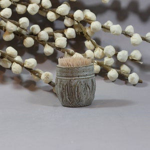 May include: A small, grey ceramic toothpick holder with a textured design. The holder is filled with toothpicks and is sitting on a light grey surface.
