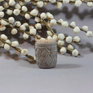 May include: A grey ceramic toothpick holder with a decorative pattern. The holder is filled with toothpicks and is sitting on a light grey surface.
