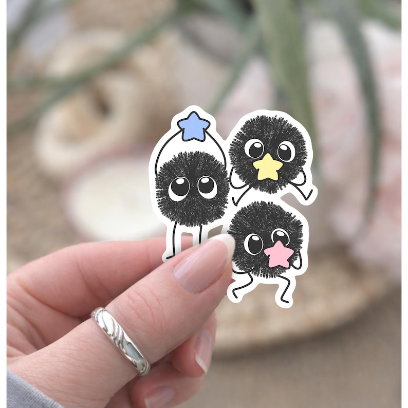 May include: A sticker featuring three cartoon characters with black, fluffy bodies and large, round eyes. Each character holds a star in a different colour: blue, yellow, and pink. The sticker is held by a hand.