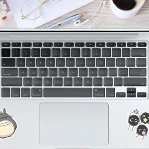 May include: A laptop with a black keyboard and a white trackpad. Stickers of a Totoro character and three soot sprites are on the laptop. A pen, glasses, and flowers are on the wooden surface.