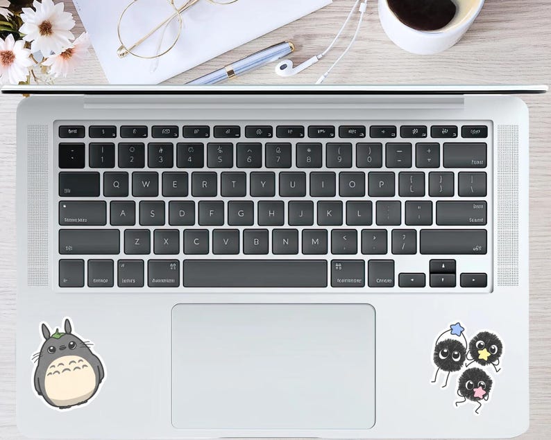 May include: A laptop with a black keyboard and a white trackpad. Stickers of a Totoro character and three soot sprites are on the laptop. A pen, glasses, and flowers are on the wooden surface.