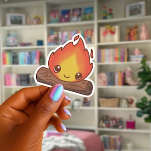 May include: A cartoon campfire sticker with a smiling face, red and yellow flames, and a brown log. The sticker is held up against a blurred background of a bookshelf filled with books.