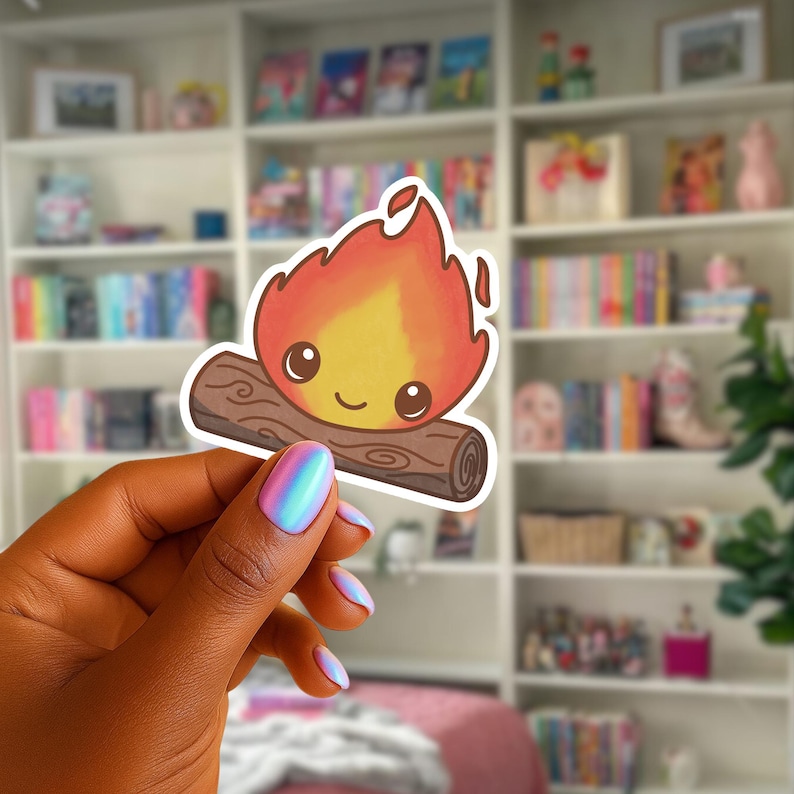 May include: A cartoon campfire sticker with a smiling face, red and yellow flames, and a brown log. The sticker is held up against a blurred background of a bookshelf filled with books.