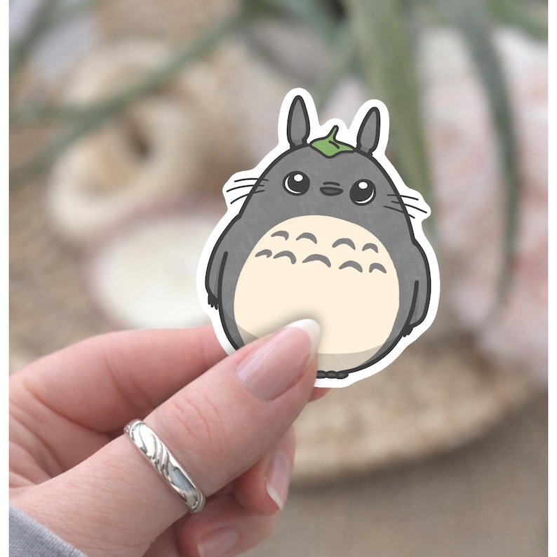 May include: A sticker depicting a cartoon Totoro character. The design shows a grey, round creature with a cream-coloured belly, large eyes, and a leaf on its head. The sticker is held by a hand with a silver ring.