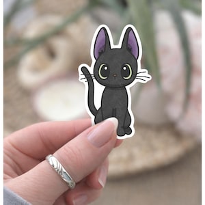 May include: A black cat sticker with large, round eyes and purple-lined ears. The cat is sitting upright with its tail curved. The sticker has a white border and is held by a hand with a silver ring.