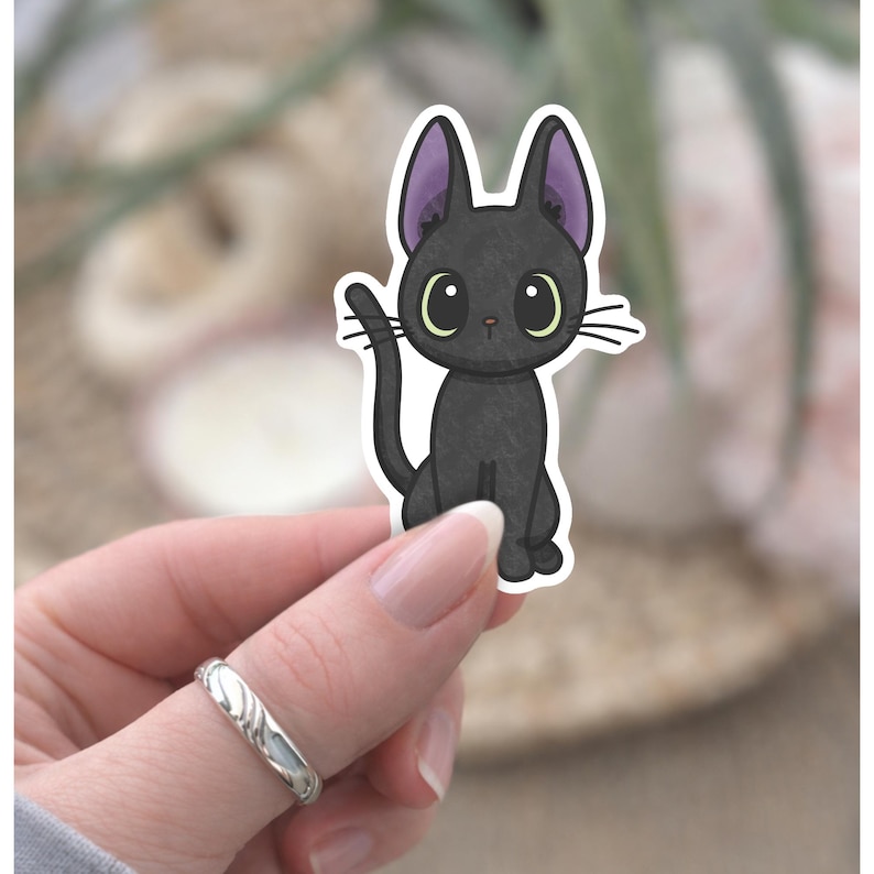 May include: A black cat sticker with large, round eyes and purple-lined ears. The cat is sitting upright with its tail curved. The sticker has a white border and is held by a hand with a silver ring.