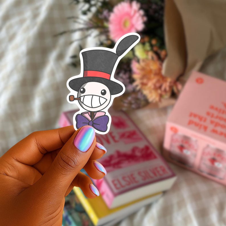 May include: A sticker depicting a cartoon character wearing a top hat, bow tie, and smoking a pipe. The character has a smiling face. The sticker is held in front of a bouquet of flowers and a book.