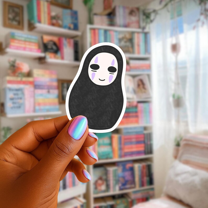 May include: A black sticker of a Spirited Away character, with a white face and purple details. The sticker is held in front of a bookshelf with a variety of colourful books.