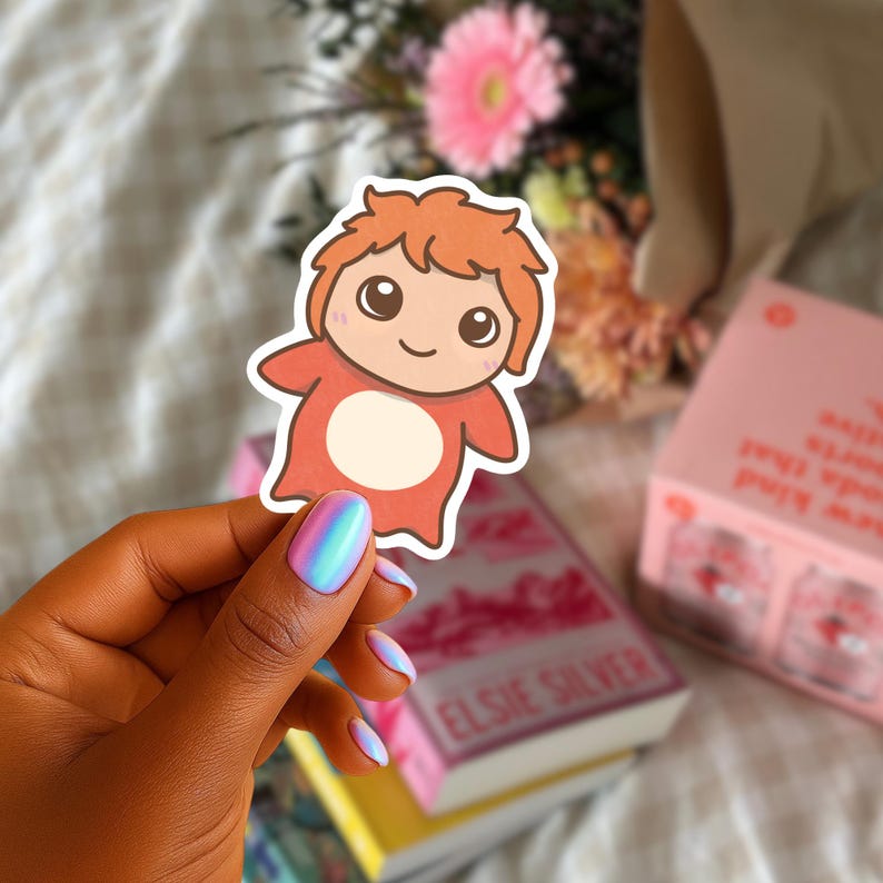 May include: A sticker of a cartoon character with orange hair and a red outfit. The character has a white belly and a smiling face. The sticker is held in front of a blurred background of flowers and books.