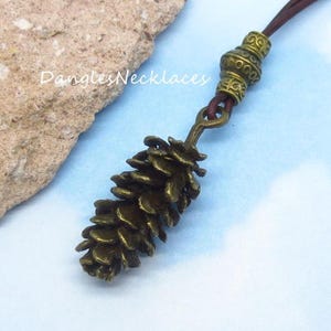 May include: A bronze-toned metal pinecone pendant hangs from a dark brown leather cord. The necklace features a decorative, etched metal bead above the pinecone charm. The text DanglesNecklaces is visible in white script on the left side of the image.