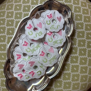 Puede incluir: Una bandeja plateada contiene varios posavasos blancos con forma de flor. Cada posavasos presenta un corazón rosa, una herradura verde e iniciales bordadas en verde. También hay una mariposa rosa bordada en cada posavasos.