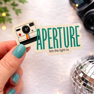 May include: Aperture sticker featuring a retro camera illustration with a rainbow stripe. The word "APERTURE" is in teal, with the phrase "lets the light in." The sticker is held by a hand with teal nail polish, with a disco ball in the background.