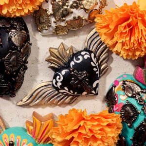 May include: A black heart-shaped ornament featuring white floral scrollwork, a central dark metal cross, and gold-toned wings and rays. The piece is surrounded by bright orange artificial marigold flowers, creating a vibrant contrast.