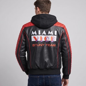May include: A black leather jacket with red quilted shoulder and sleeve panels, featuring a black hood and the text MIAMI VICE STUNT TEAM printed in white and orange on the back. The jacket has a ribbed black hem and cuffs and red piping details.