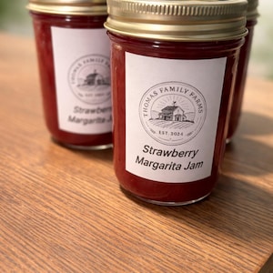 Könnte beinhalten: Mehrere Gläser mit roter Marmelade, jeweils mit einem goldenen Deckel und einem weißen Etikett. Auf dem Etikett steht "Thomas Family Farms" und "Strawberry Margarita Jam". Die Gläser stehen auf einer Holzoberfläche.