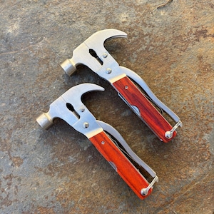 May include: Two multi-tool hammers with stainless steel heads and red wooden handles. The hammers have a claw, pliers, and a small knife. The tools are designed for various tasks and are suitable for home improvement or outdoor use.