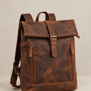 Peut inclure: Un sac à dos en cuir marron doté d'une fermeture à rabat enroulable sécurisée par une sangle verticale unique et une boucle en métal. Le sac comprend deux poches zippées verticales sur le devant, une poignée supérieure et des bretelles réglables.